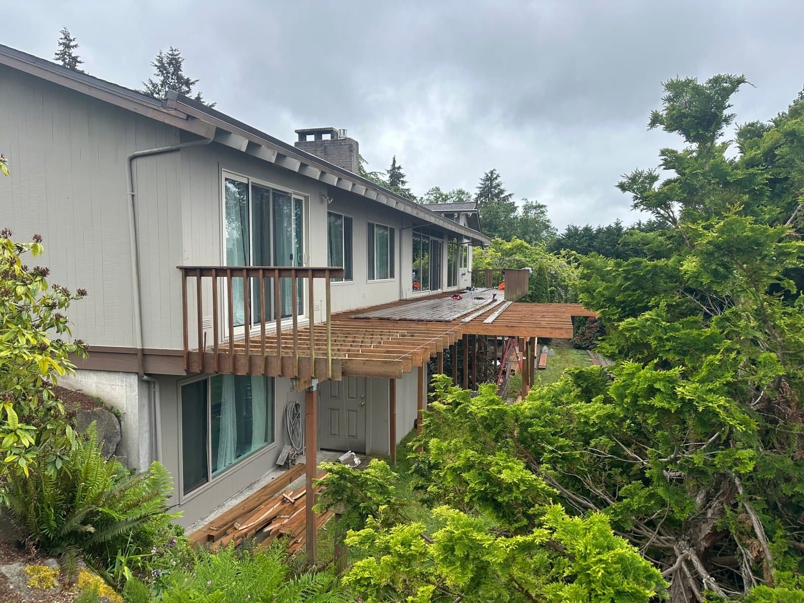 A house with a partially built wooden deck is surrounded by green trees on a cloudy day.