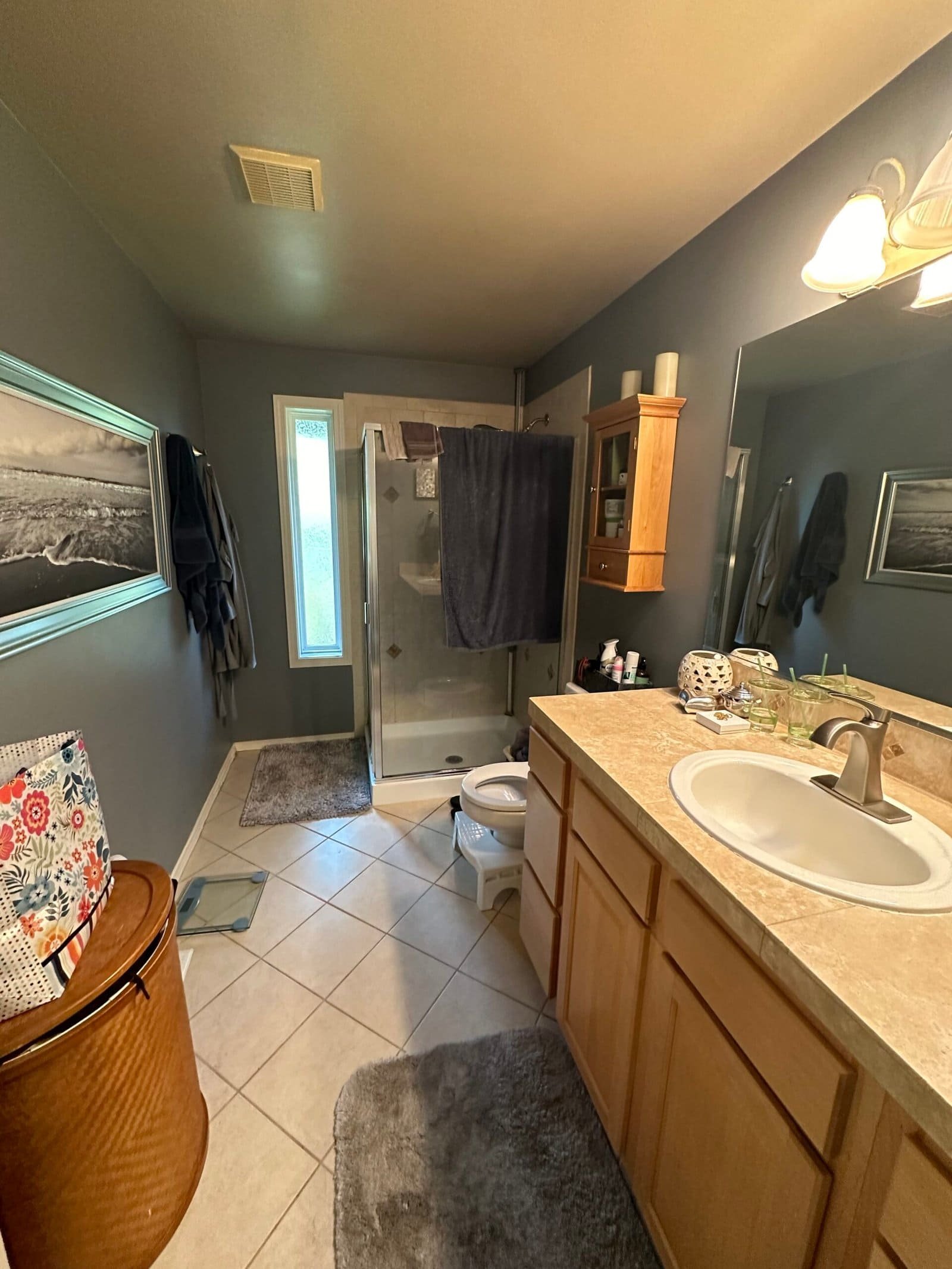 A modern bathroom with tiled flooring features a sink, mirror, shower, towels, and laundry basket.
