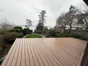 A wet wooden deck overlooks a garden, trees, and water, ideal for outdoor living on cloudy days.