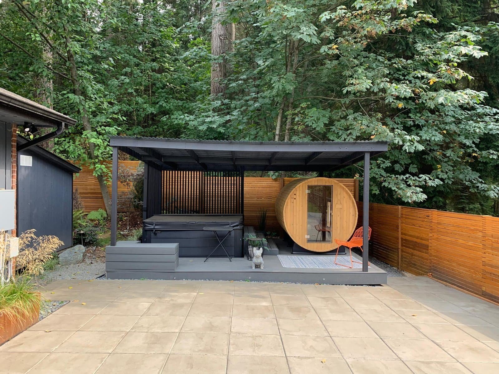 A modern backyard features a covered hot tub, barrel sauna, patio seating, and wooden fence among trees.