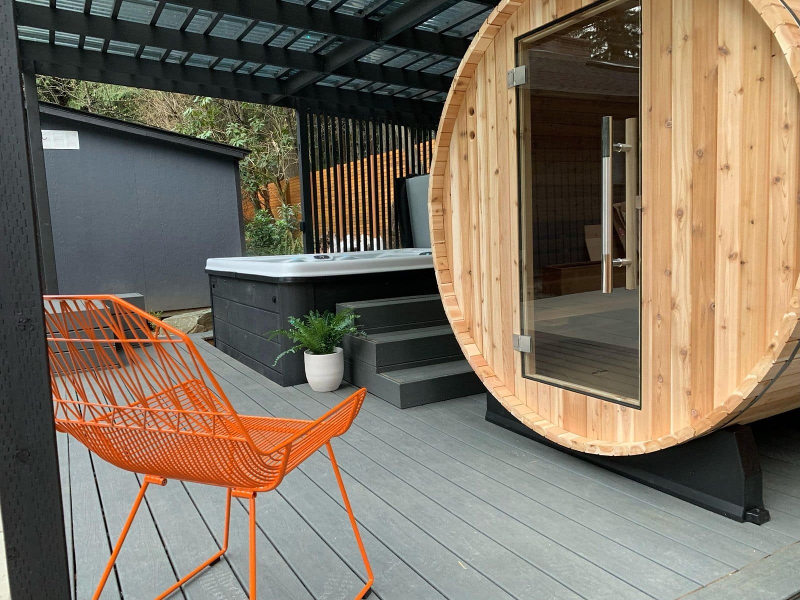 A modern outdoor deck features an orange chair, a hot tub, and a round wooden sauna for relaxation.
