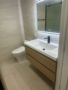 Modern bathroom featuring a lit mirror, white sink, wood vanity, toilet, and light tile walls and floor.