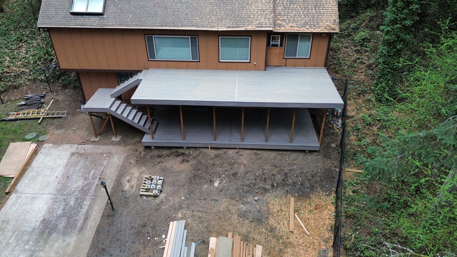 A house featuring a covered wooden deck with stairs, surrounded by trees, dirt, and visible construction materials.