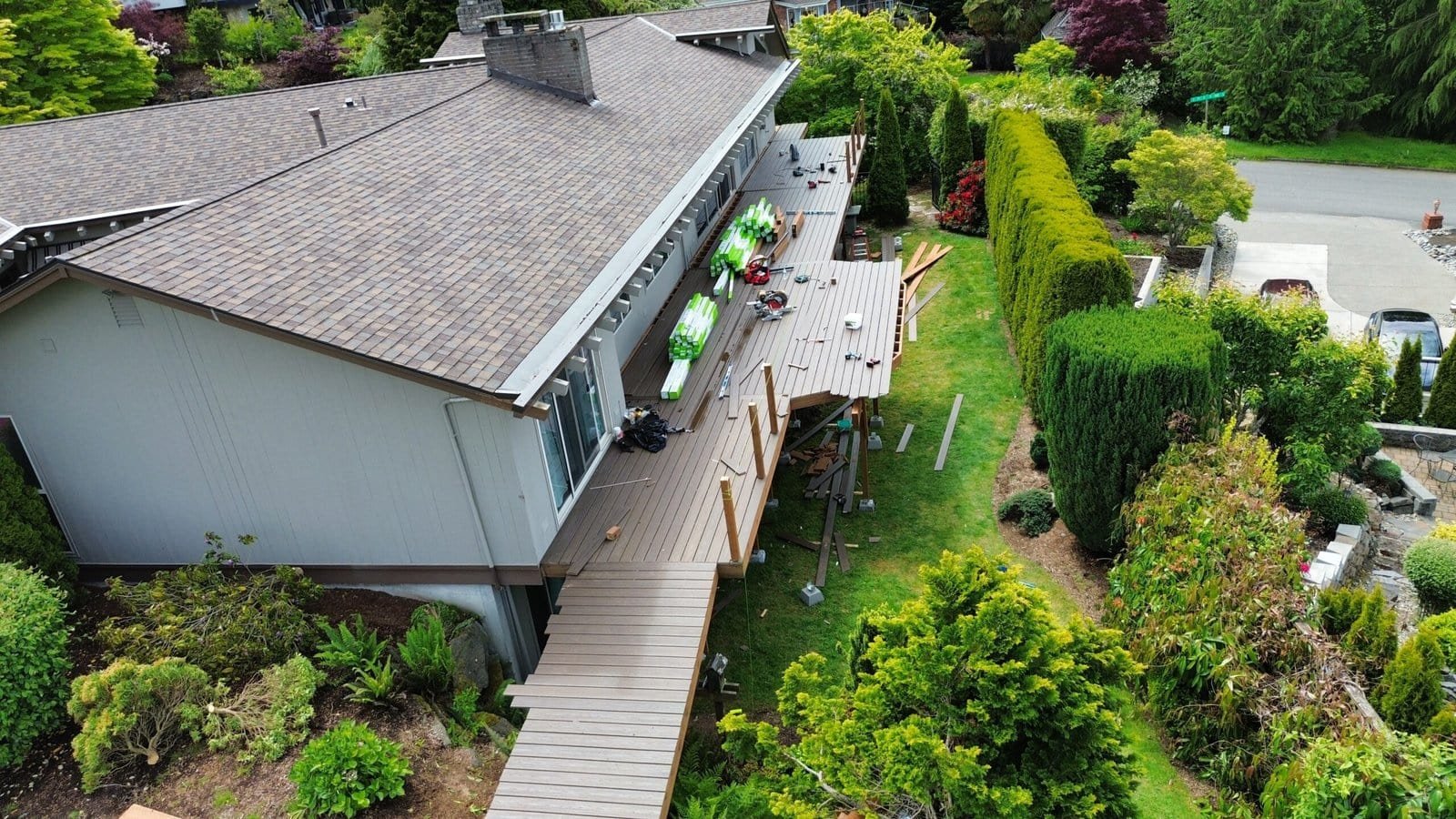 Aerial view of a backyard deck under construction, attached to a house with green trees and shrubs nearby.