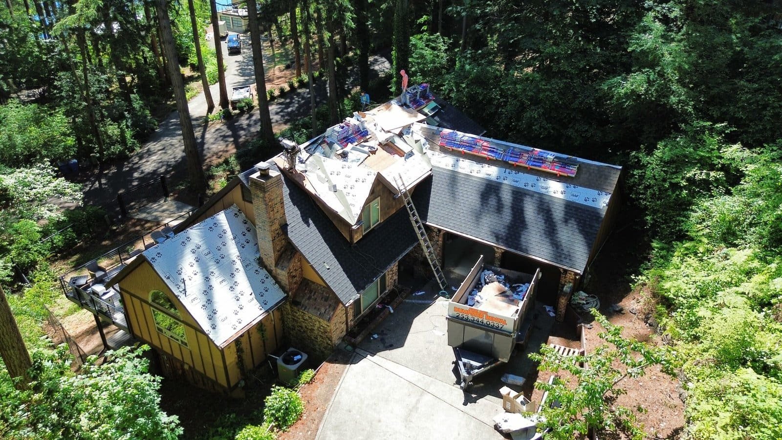 Aerial view of roofing contractors installing a new roof on a home surrounded by lush trees and greenery.