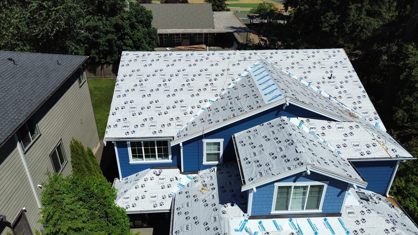 A blue house with roofing underlayment is viewed from above on a sunny day, highlighting roof details.