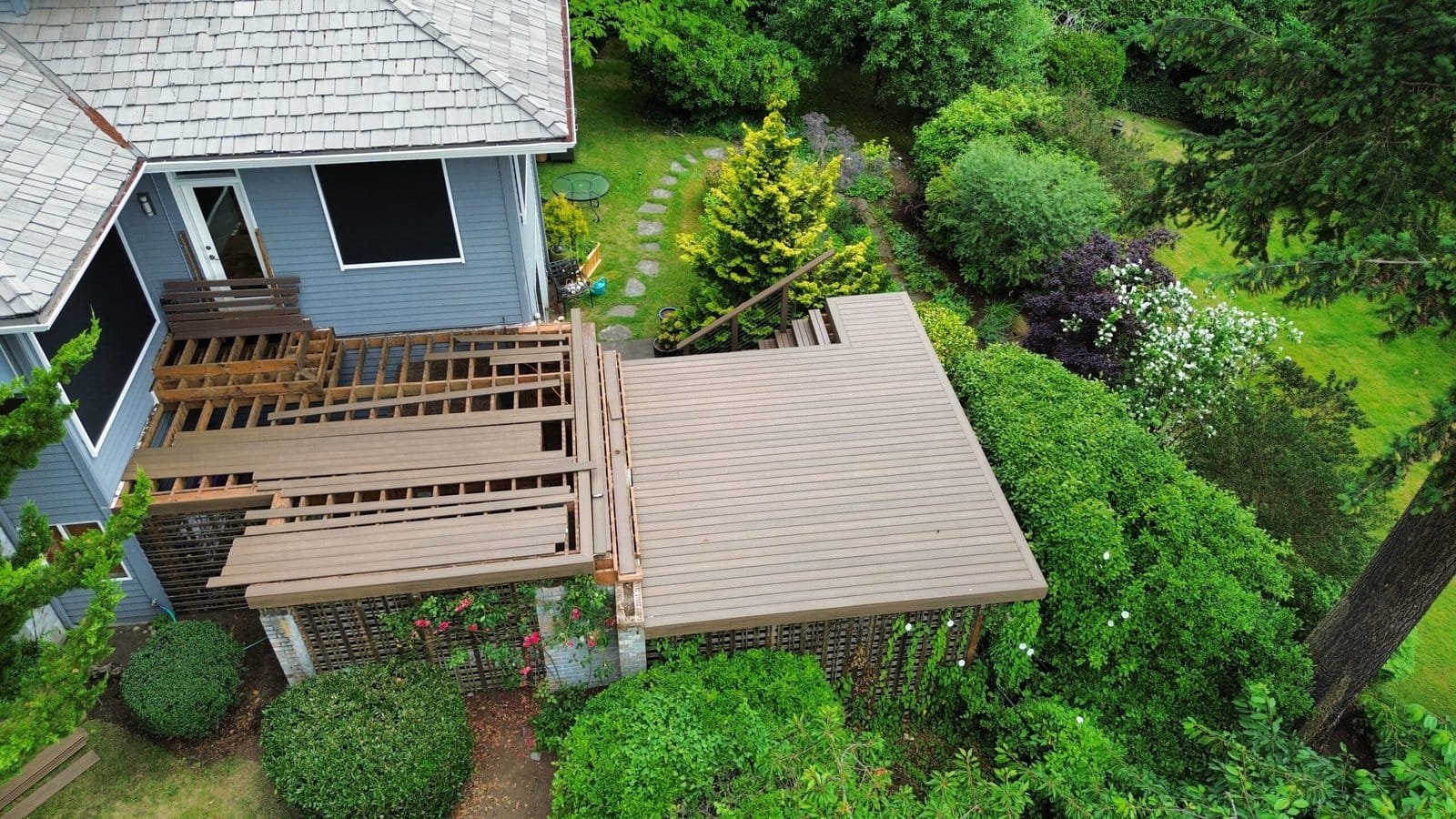 A house features a partially built wooden deck, surrounded by lush green trees and bushes in the yard.