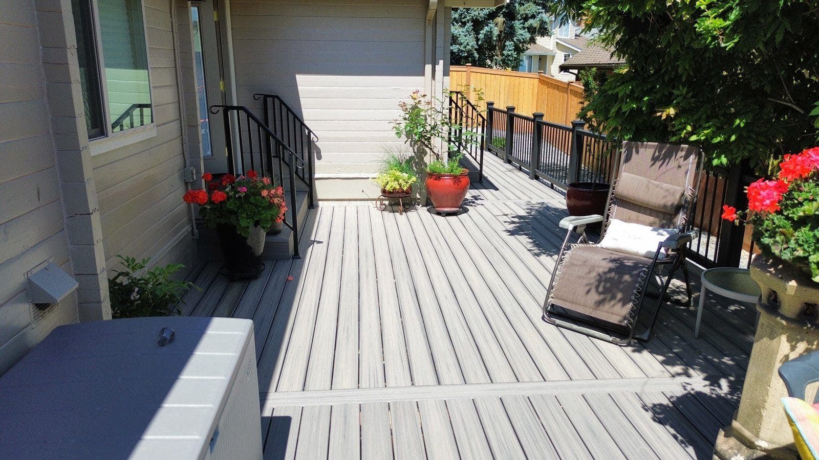A sunny patio with potted plants, lounge chair, railing, and light-colored decking beside a modern house.