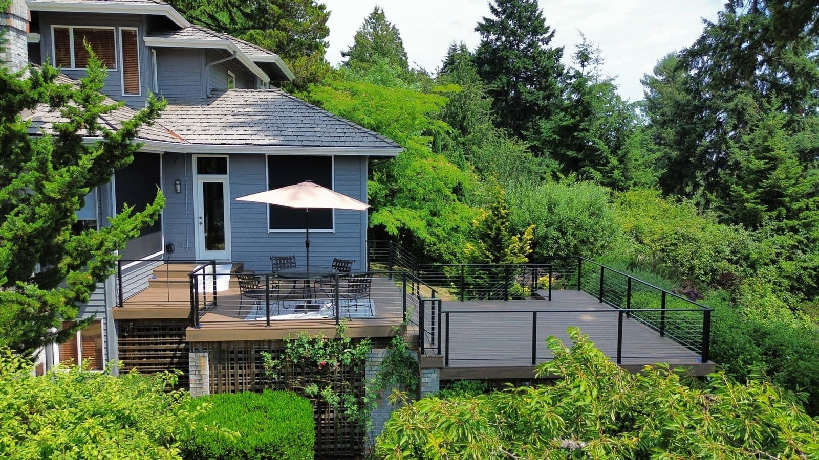 Gray house features a spacious deck with patio furniture and umbrella, set among lush green trees and plants.