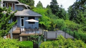 Gray house with a spacious deck, outdoor table, chairs, and umbrella set amid lush green trees.