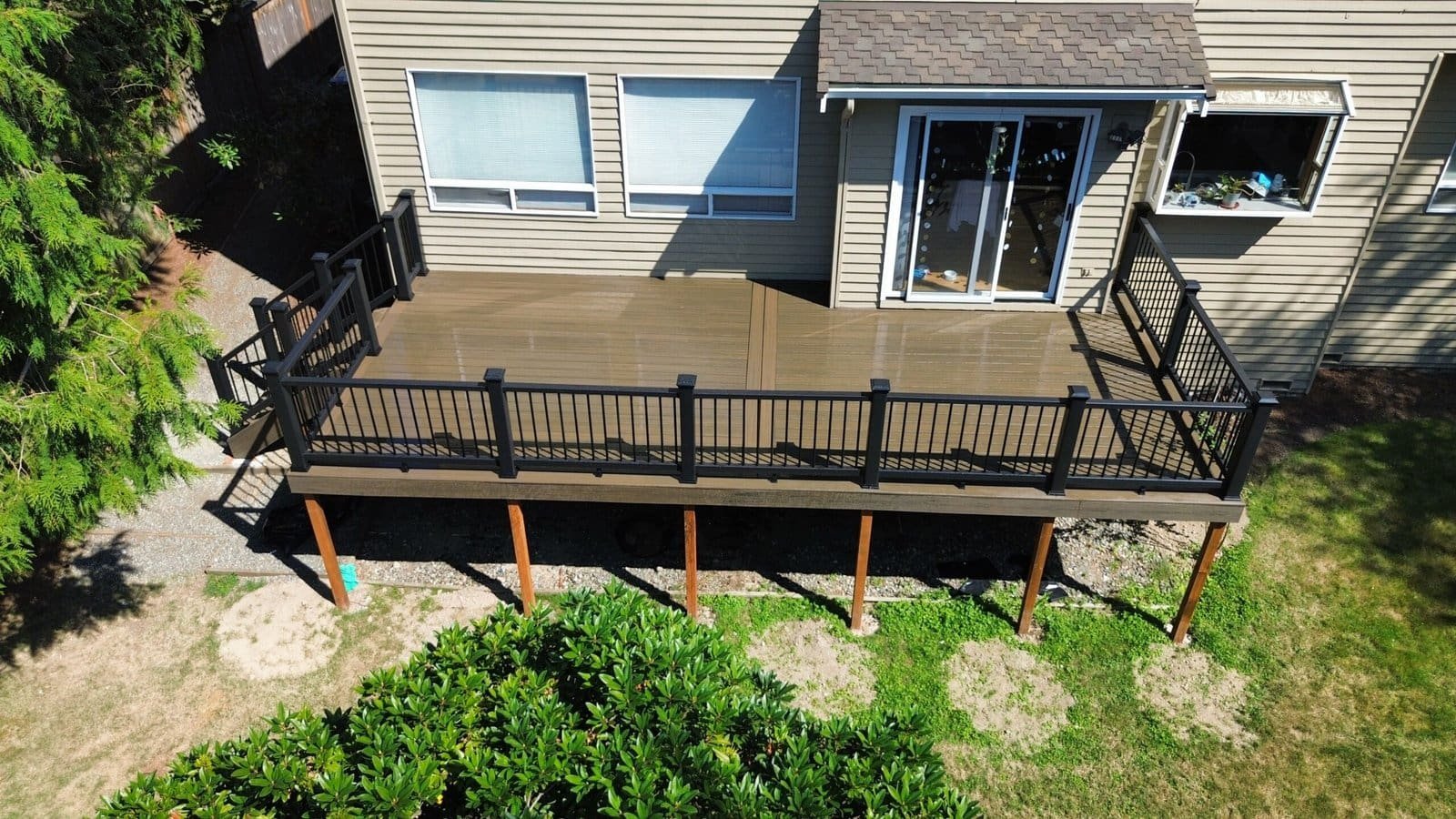 A raised wooden deck with black railings connects to a beige house featuring large windows and sliding door.