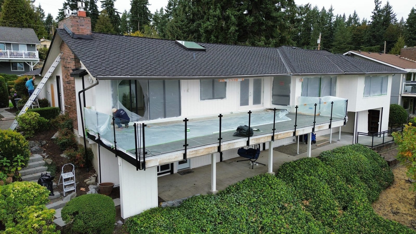 A person installs glass balcony railing on a white house with sloped roof, surrounded by greenery.