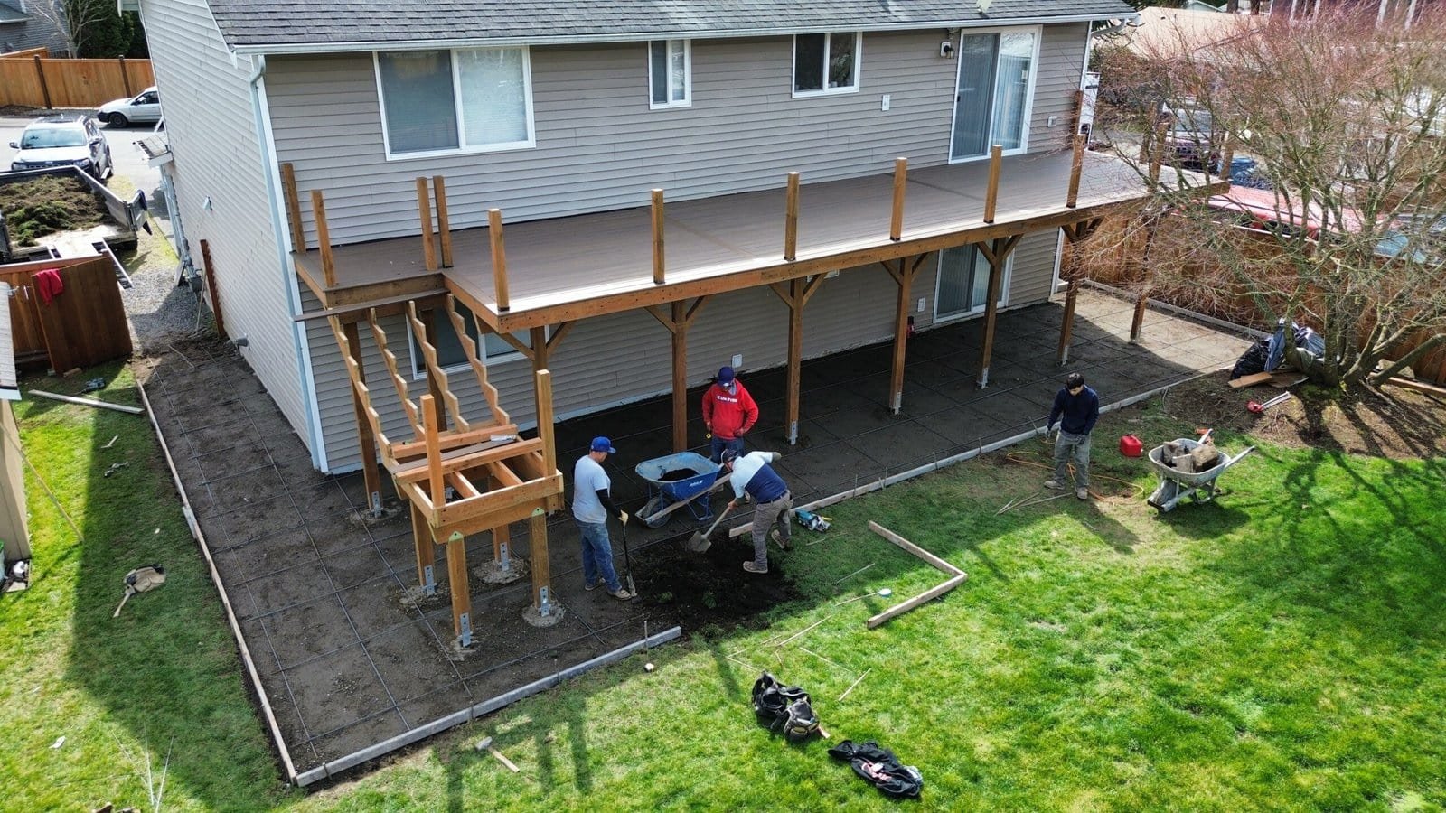 Four people are working on a backyard patio under a raised wooden deck by a two-story family house.