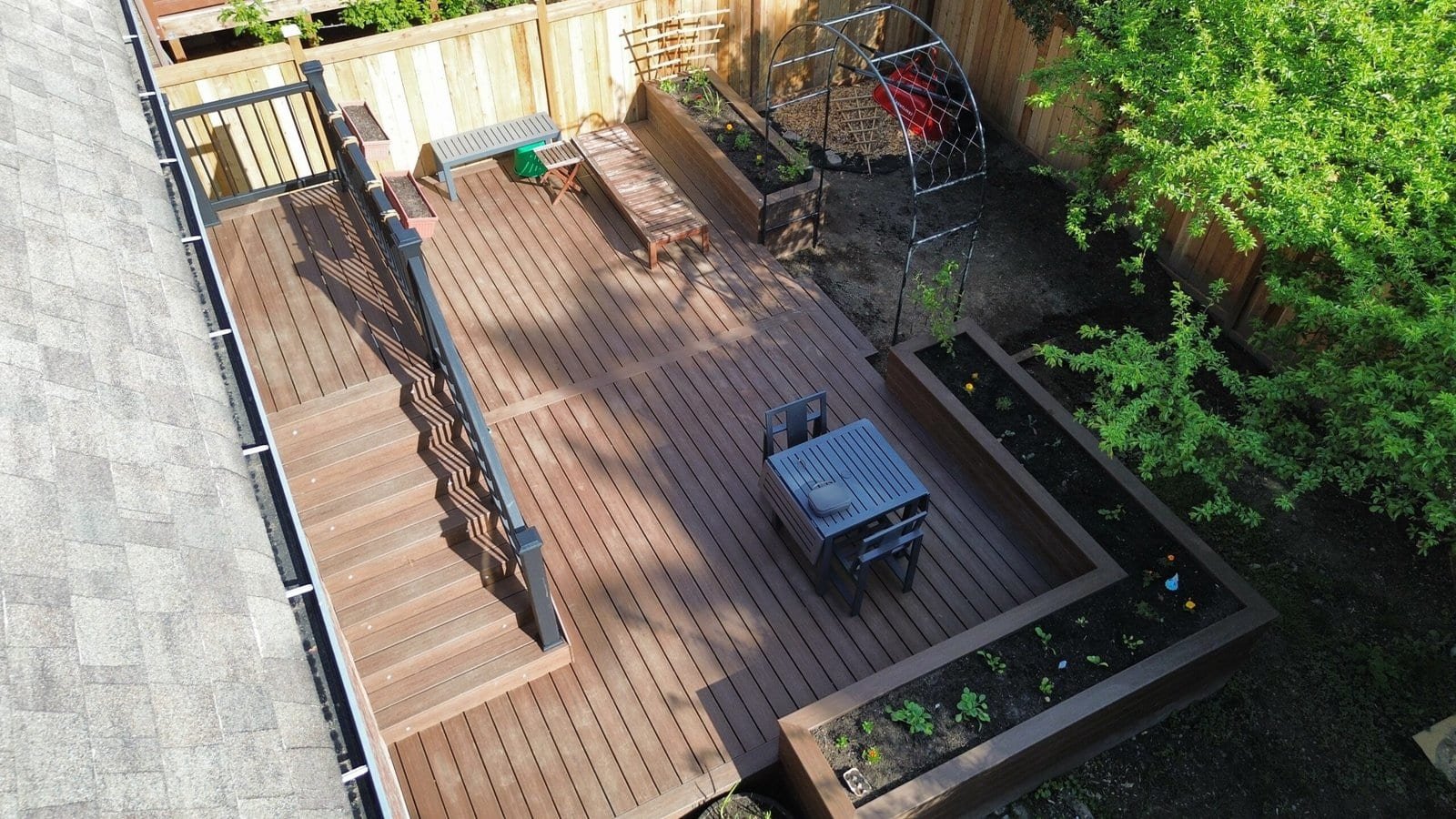 A wooden backyard deck features outdoor seating, a small table with chairs, garden beds, and a leafy green tree.