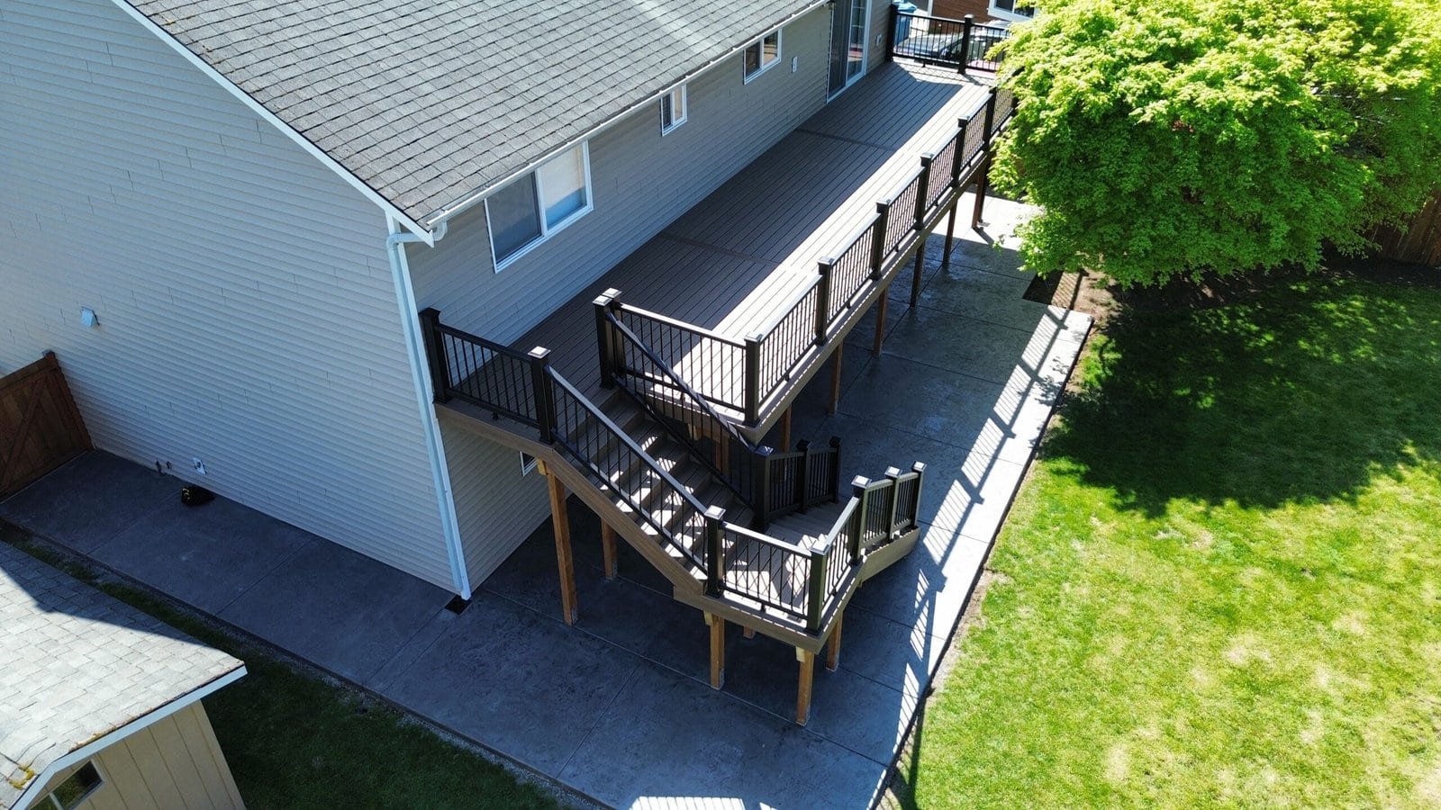 Aerial view of a house features a raised deck, stairway, concrete patio, green lawn, and a mature tree.