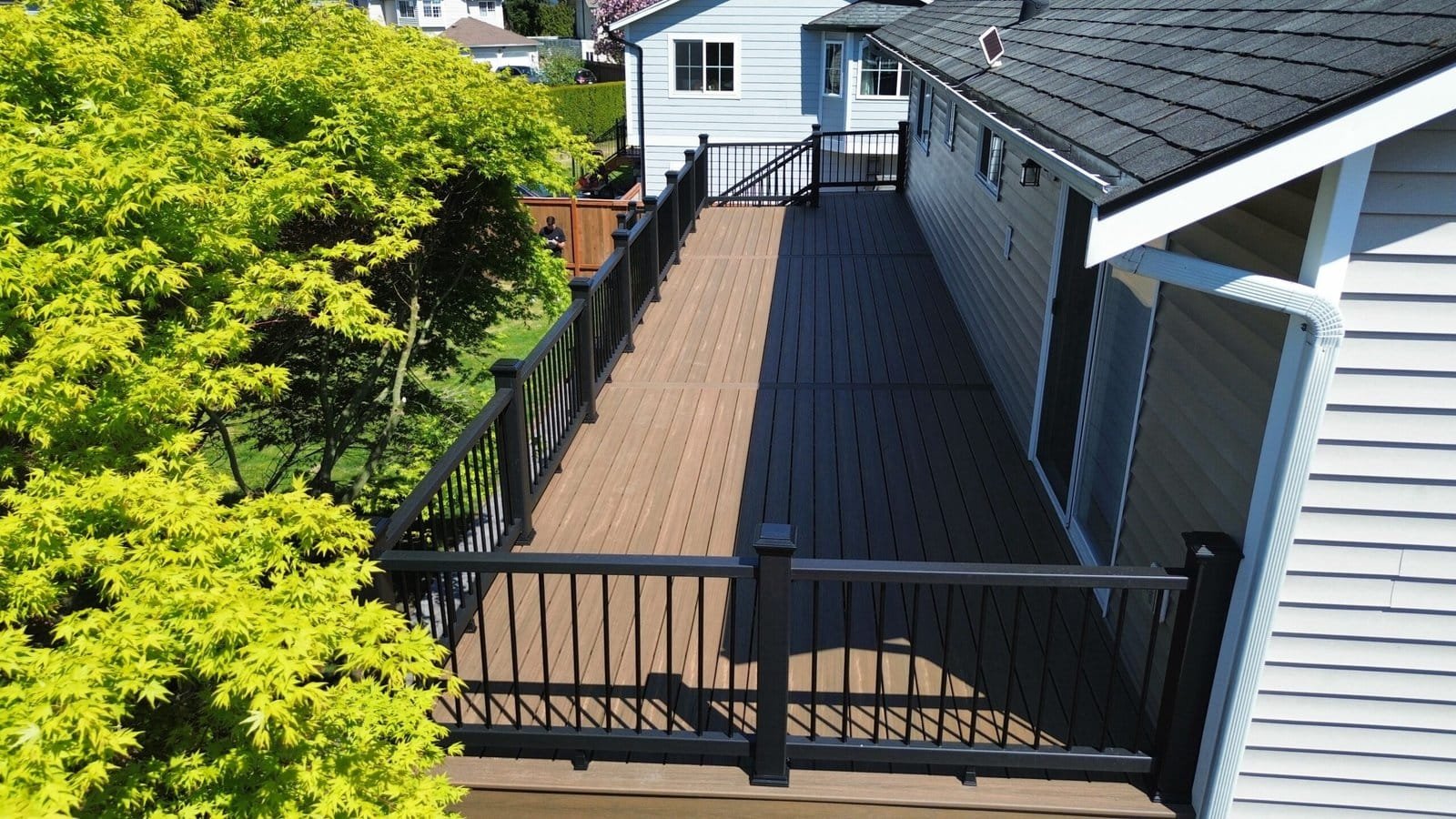 A large wooden deck with black railings is attached to a light-colored house, surrounded by green trees.
