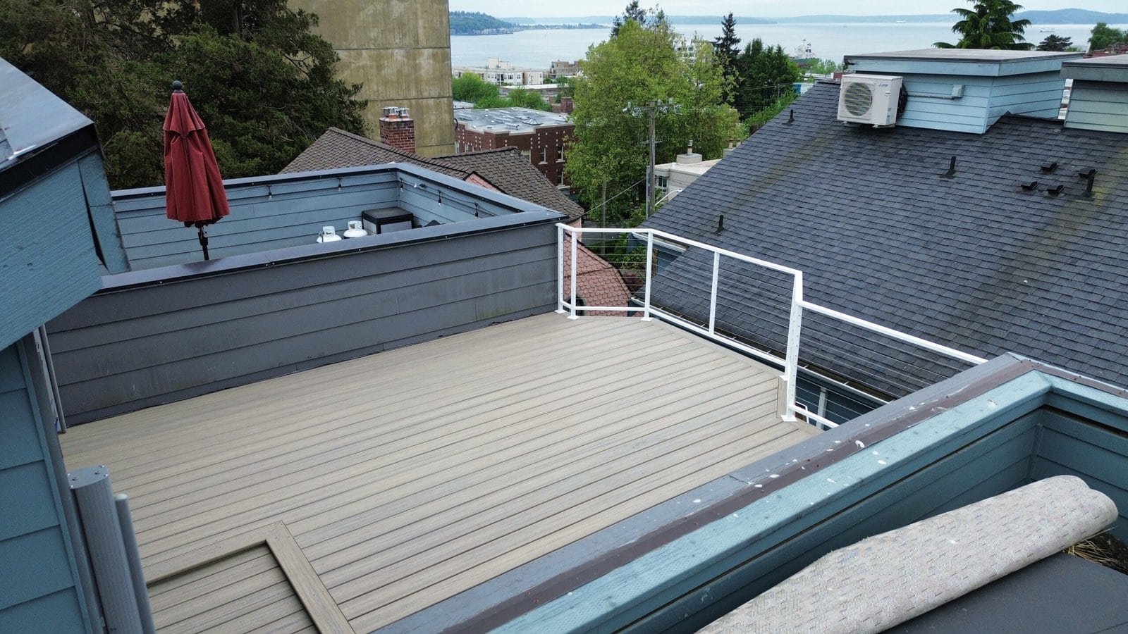 Rooftop deck with light wood flooring, white railing, red umbrella, and a scenic water view beyond trees.