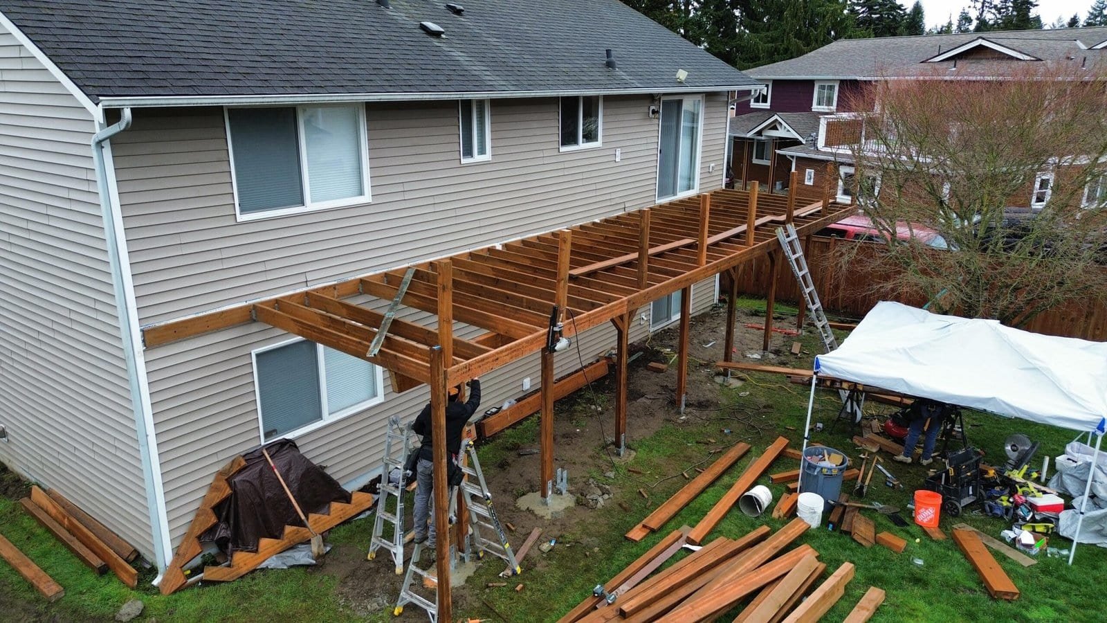 A new wooden deck under construction behind a two-story house features tools and materials scattered nearby.