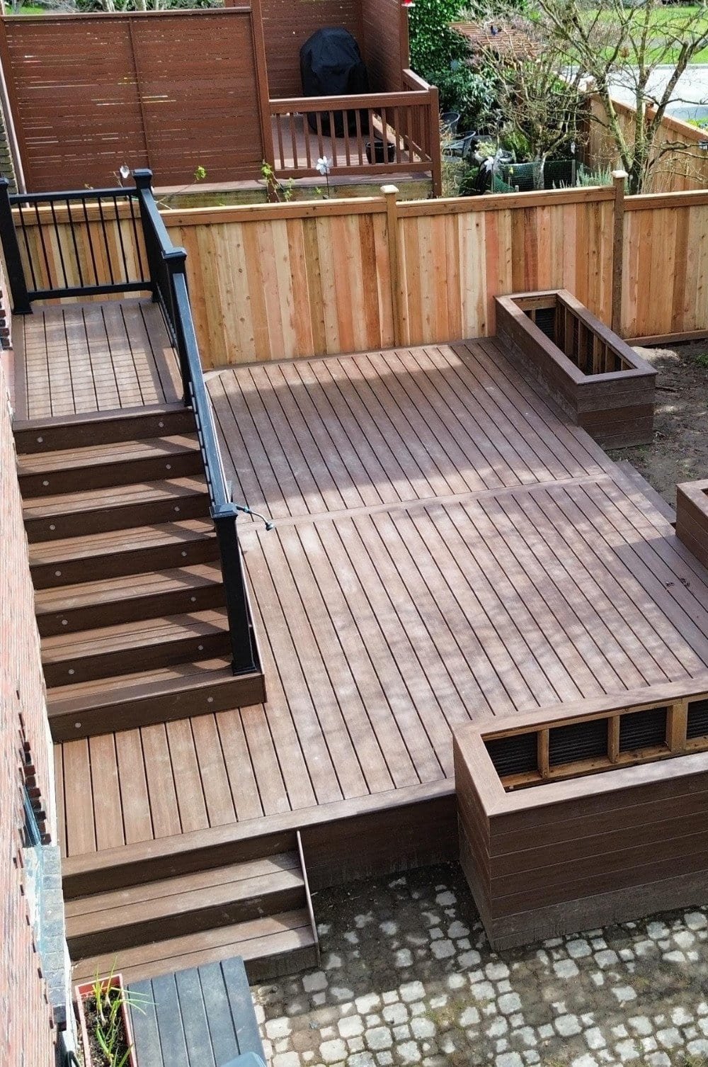 A multi-level wooden deck with stairs, railing, and built-in planters is set in a fenced backyard.