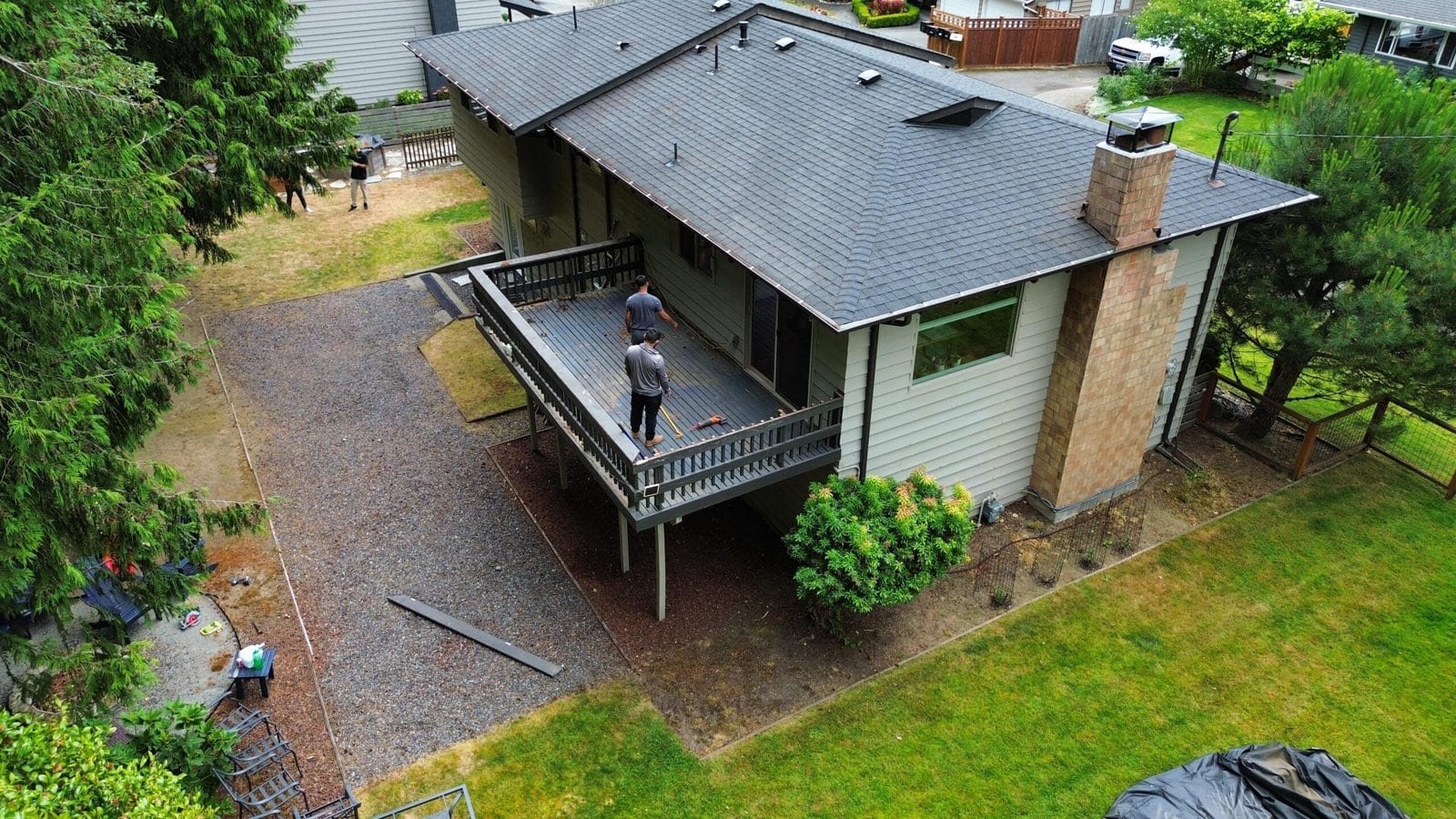 Two people build a wooden deck on a beige house with a gray roof, as viewed from above.