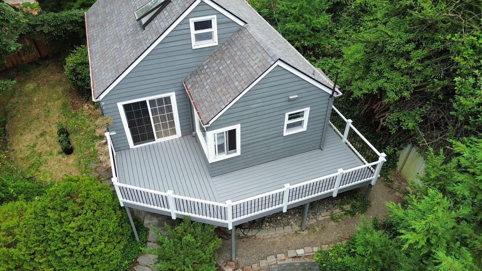 Gray house with white trim and a large elevated deck sits amid trees and greenery in this peaceful scene.
