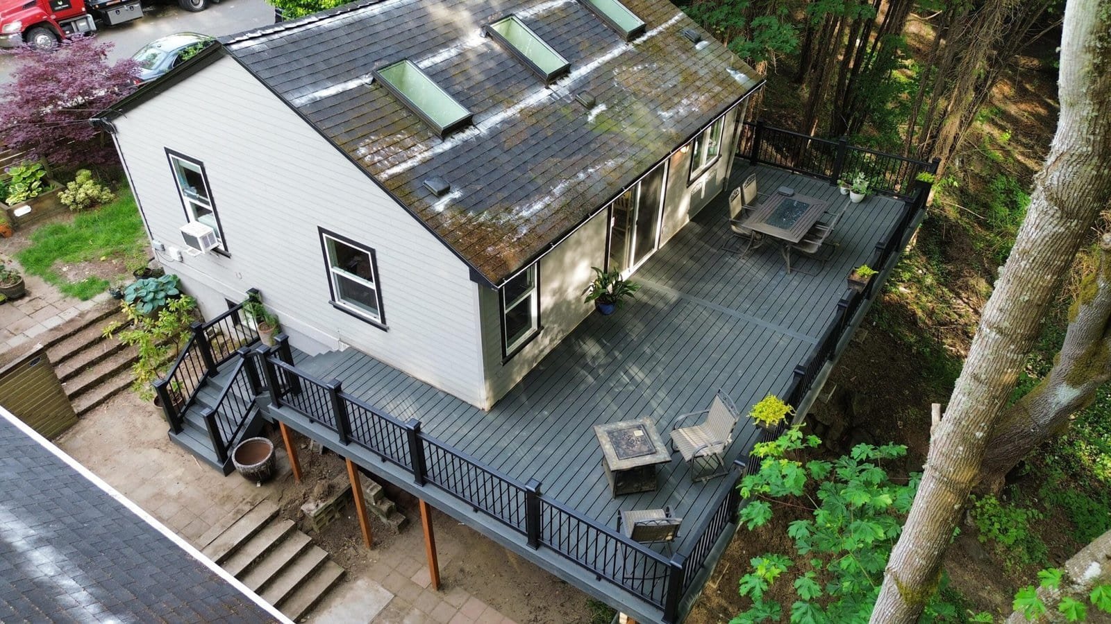 Aerial view of a house with skylights, a large gray deck, outdoor seating, and surrounded by trees.
