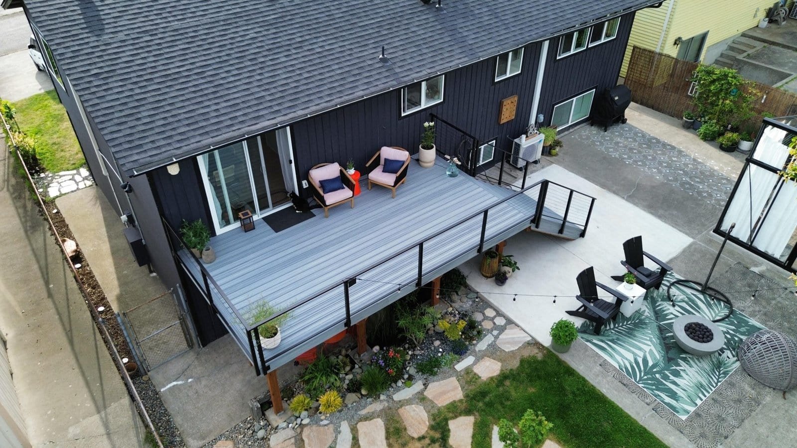 A modern house features a raised deck with outdoor seating and a cozy fire pit area with chairs and rug.