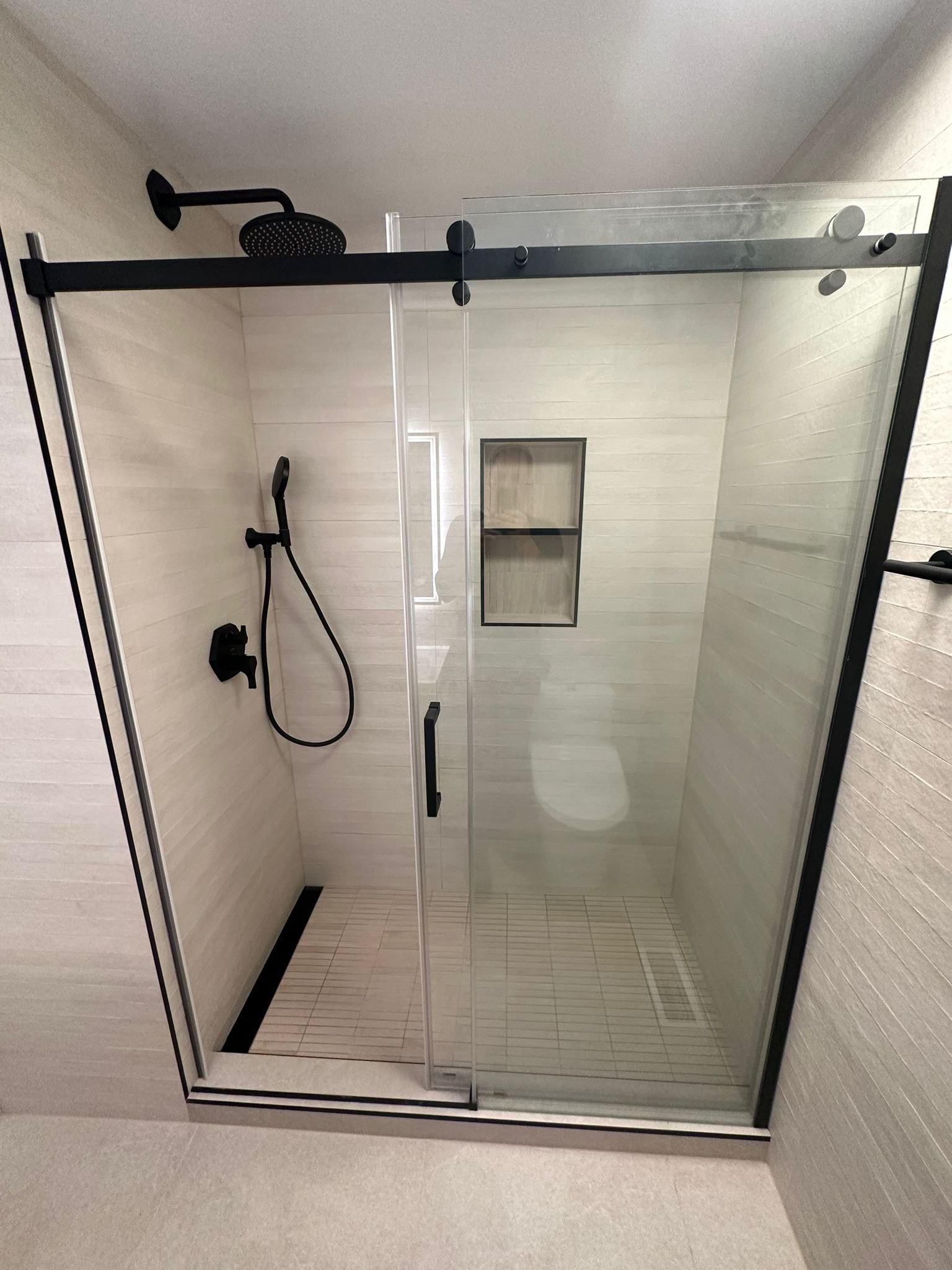 Modern shower with glass doors, black fixtures, a rainfall showerhead, and built-in wall shelf shown.
