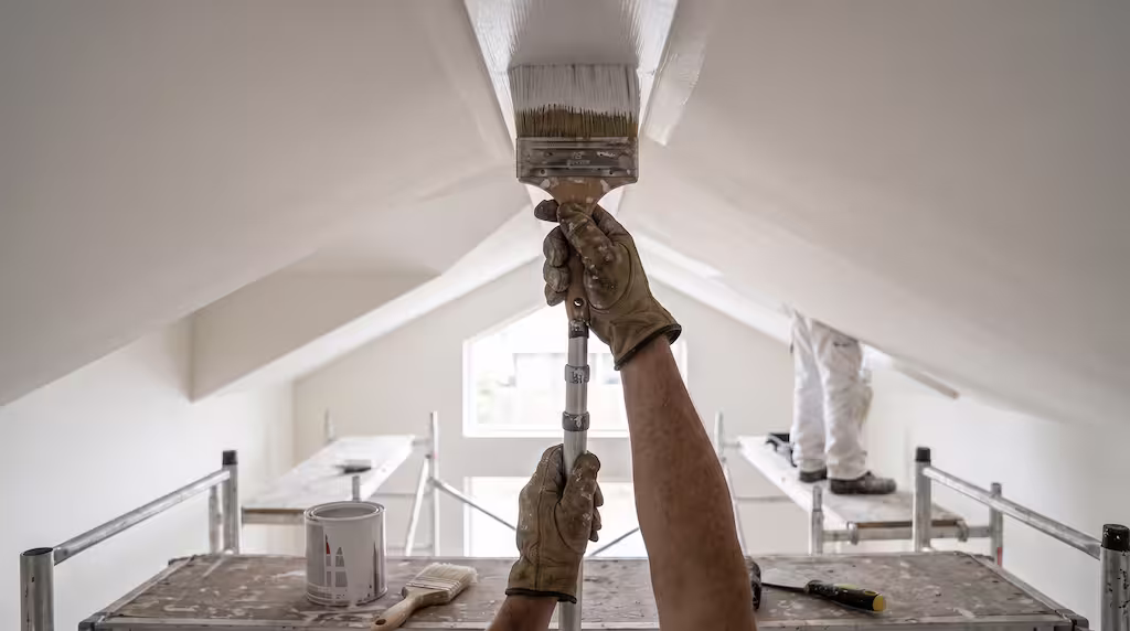 A person in leather gloves uses an extension pole and brush to paint a white ceiling.