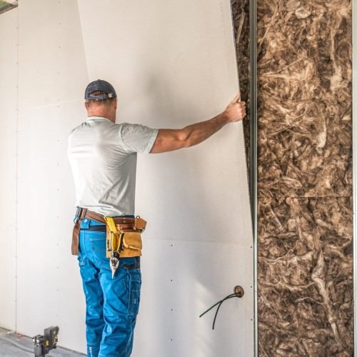 A drywall contractor installs drywall in a room with exposed insulation, wearing a tool belt around his waist.
