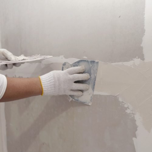 A drywall contractor in white gloves smooths plaster on a wall seam with a putty knife for finishing.