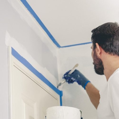 Man performing interior painting uses masking tape for crisp edges while painting a wall white near ceiling.