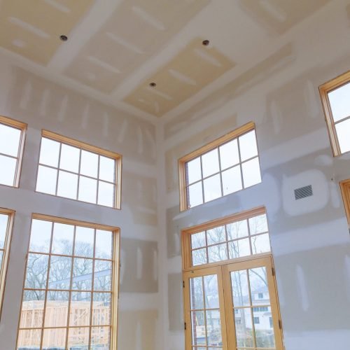 Interior room with unfinished drywall by a skilled contractor, large windows show trees and nearby houses.