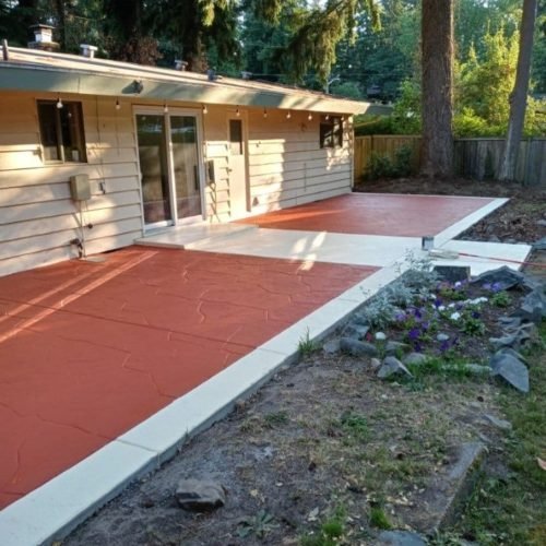A fenced backyard features a house with a red and white concrete patio, garden bed, trees, and driveway.