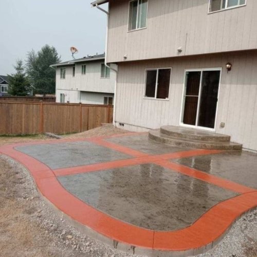 A wet concrete patio with a red border and X pattern sits by the driveway behind a beige two-story house.