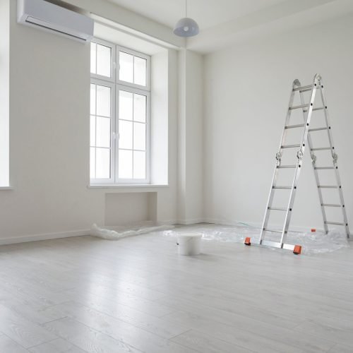 Empty room ready for interior painting with large windows, a metal ladder, paint bucket, and plastic sheet.