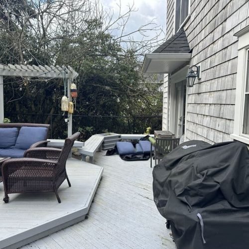 A wooden deck featuring a barbecue grill and several chairs arranged for outdoor cooking and dining. The deck surface consists of neatly aligned wooden planks, and sunlight casts natural shadows across the seating and grill area. The scene depicts a cozy and functional outdoor space for gatherings and meals