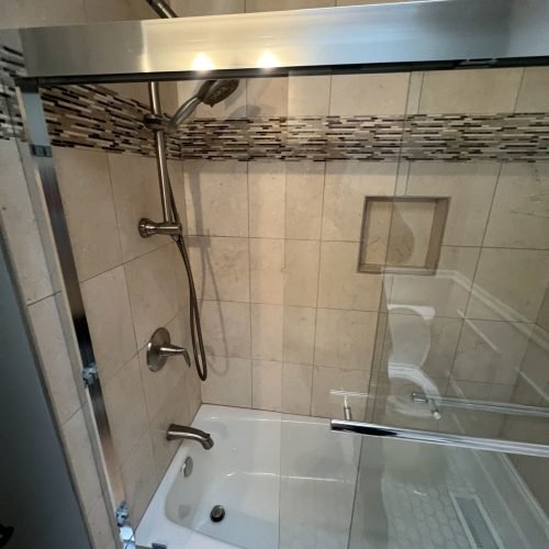 A bathroom featuring a shower enclosed with a glass door and a toilet positioned nearby. The shower area has tiled walls, and the bathroom is clean, organized, and well-lit. The layout is functional, combining bathing and toilet facilities in a compact space.