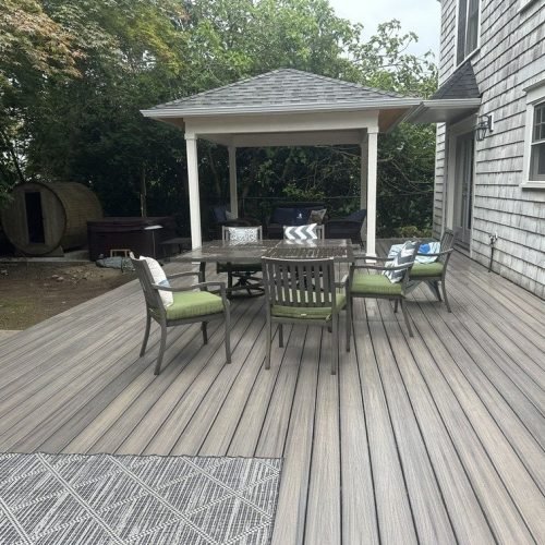 A wooden deck featuring a gazebo with a roof, providing shade for several chairs arranged underneath for relaxation or outdoor dining. The deck has neatly arranged wooden planks, and sunlight casts soft shadows across the seating area. The scene creates an inviting and comfortable outdoor space.