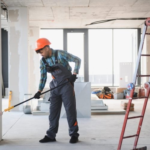Construction worker in hard hat uses a roller to paint an unfinished room, with a ladder nearby.