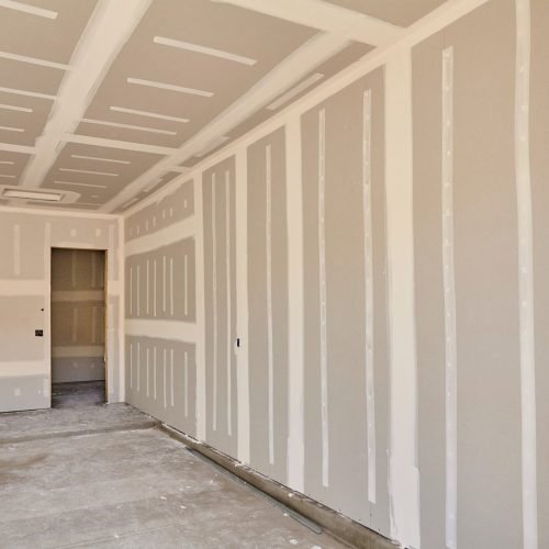 A room under construction reveals a drywall contractor finishing unfinished panels and joint compound.