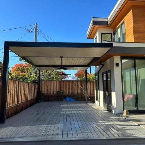 A modern house with a spacious wooden deck and a black-framed pergola. The deck is empty, bordered by a wooden fence, with clear blue sky and autumn trees in the background.