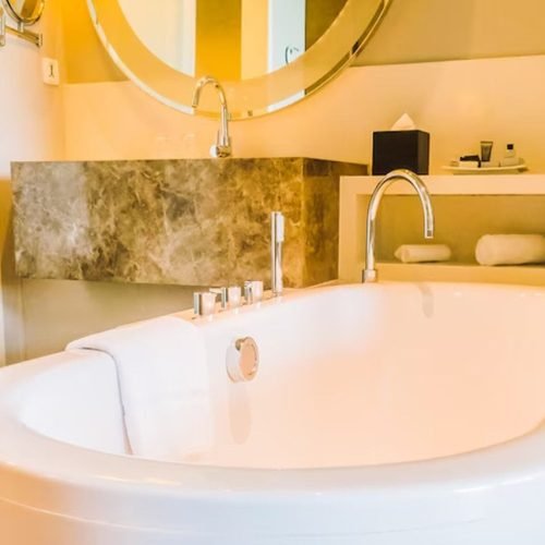 A spacious bathroom featuring a large, freestanding bathtub positioned in front of a wide wall-mounted mirror. The bathroom has light-colored tiles, modern fixtures, and a clean, minimalist design, creating a bright and luxurious atmosphere for relaxation.
