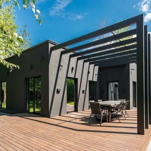 A modern black-painted house featuring wooden decking and a spacious patio area. The patio may include outdoor furniture and decorative elements, creating a stylish and functional outdoor living space. Sunlight highlights the contrast between the dark exterior and the warm wooden deck.