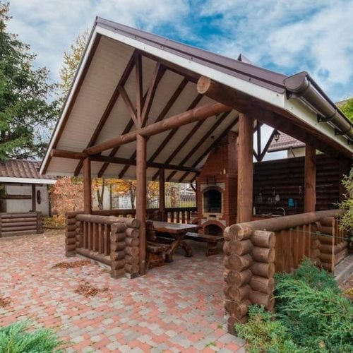 A rustic log cabin featuring a covered patio equipped with a grill. The patio has space for outdoor seating and is shaded by a roof extension. The cabin is surrounded by natural landscaping, such as trees or grass, creating a cozy and inviting outdoor area for cooking and relaxation.
