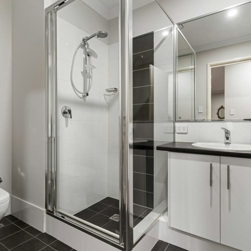 A bathroom featuring a compact shower stall, a toilet, and a sink with a countertop and mirror. The walls and floors may be tiled or painted, and the space is clean, organized, and designed for practical daily use.