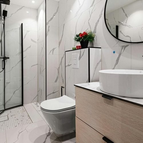 A contemporary bathroom featuring walls and flooring made of polished marble. The space includes a sink with a countertop, a toilet, and a shower or bathtub. The marble surfaces reflect light, creating a luxurious, clean, and elegant environment.